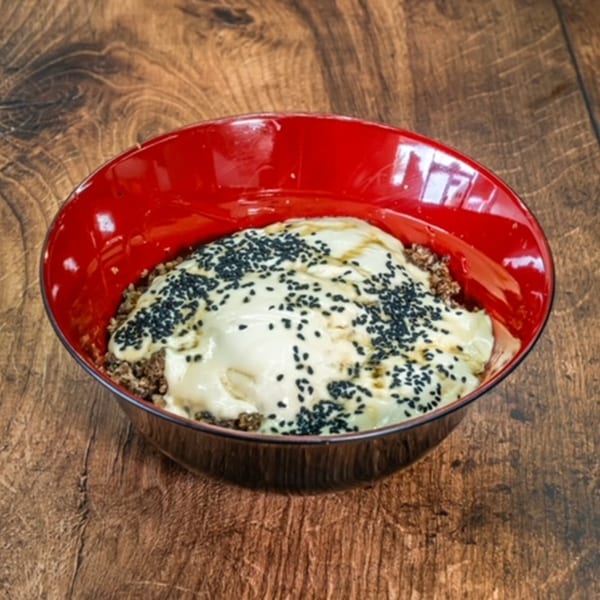 A red bowl of Middle Eastern dessert, topped with cream and black sesame seeds, on a wooden table.