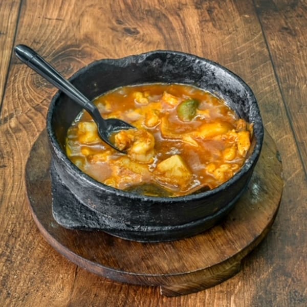 A black bowl of Bab Al-Yemen's middle-eastern stew with shrimp and vegetables, on a wooden surface.