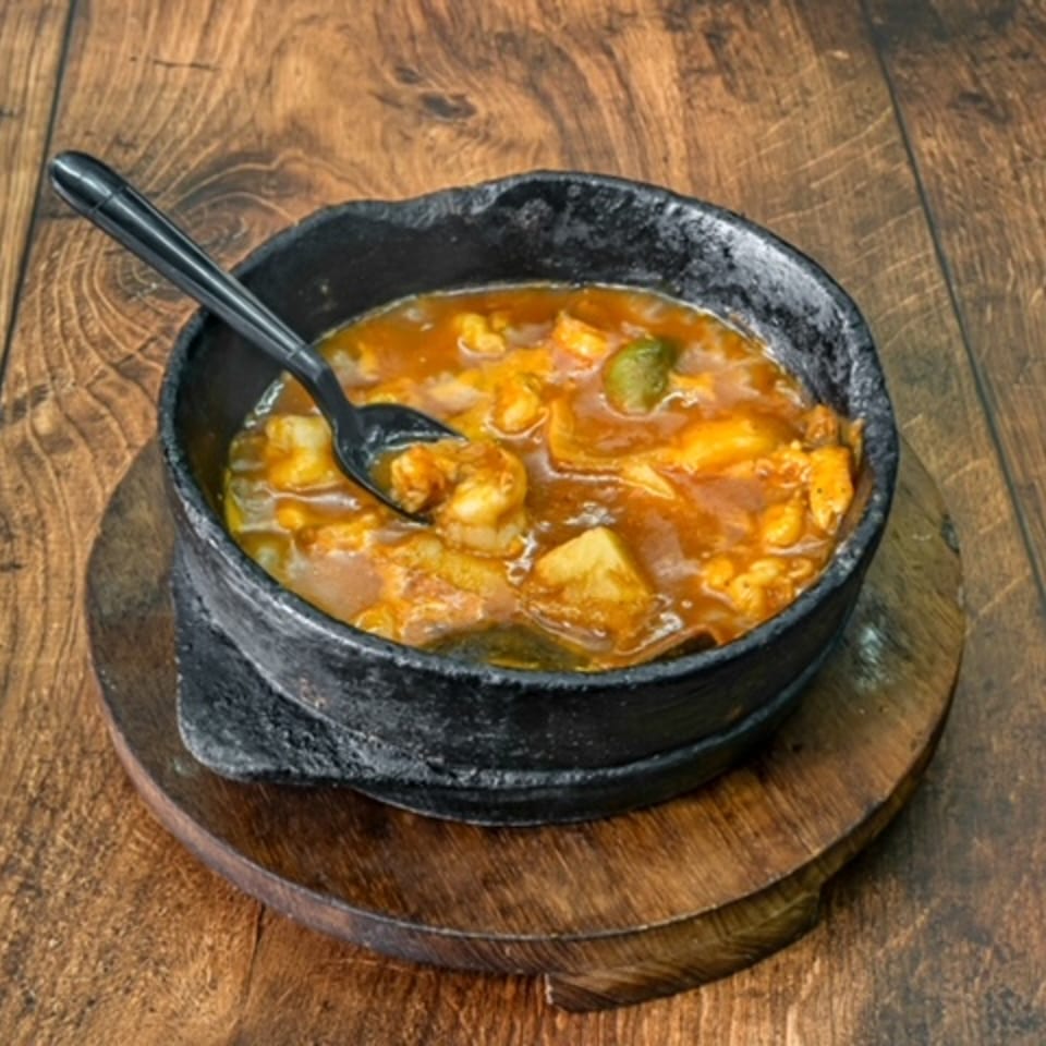 A black bowl of Bab Al-Yemen's middle-eastern stew with shrimp and vegetables, on a wooden surface.