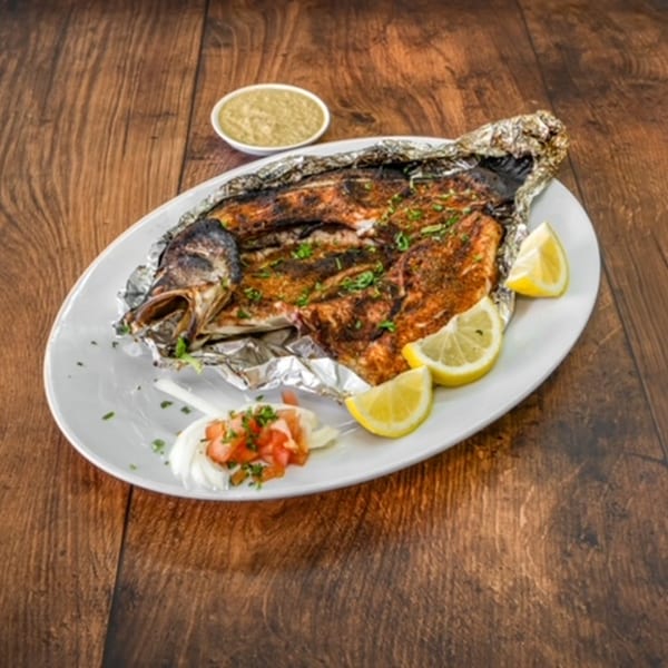 Whole grilled fish wrapped in foil, garnished with lemon and salsa, with a side of green sauce.